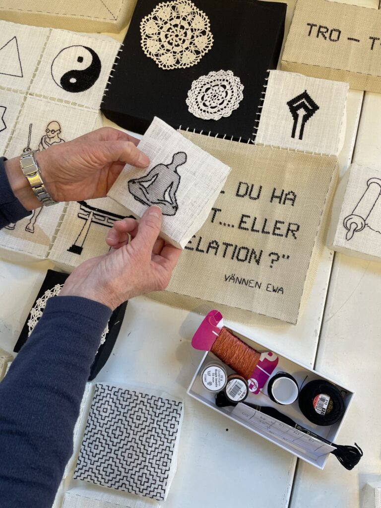 Händer håller ett textilprov framför en samling broderier, symboler och skisser på en vägg.