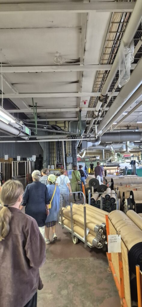 Gruppen vandrar genom en stor produktionshall med textilmaskiner hos FOV Fabrics.