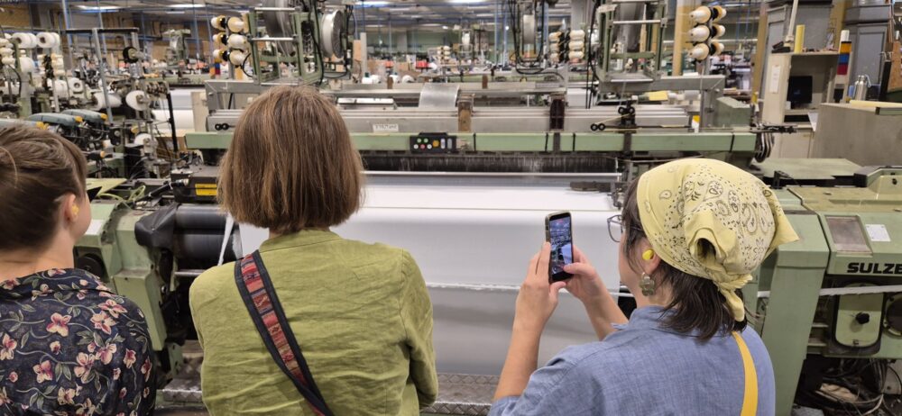 Studerande följer vävningen på nära håll hos FOV Fabrics; en person fotograferar vävmaskinen.