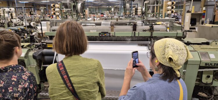 Studerande följer vävningen på nära håll hos FOV Fabrics; en person fotograferar vävmaskinen.